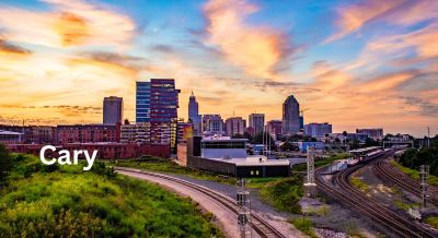 Sunset over Cary, NC, with a colorful sky, urban skyline, lush greenery, and railway tracks in the foreground.