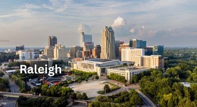 Skyline of Raleigh, NC, featuring modern high-rise buildings and lush green areas under a bright, partly cloudy sky